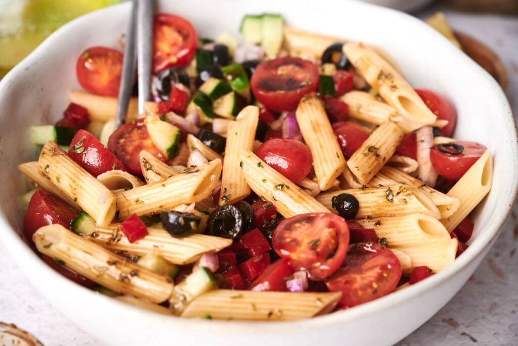 A bowl of pasta salad featuring penne, cherry tomatoes, black olives, cucumber, red onion, and fresh herbs, with a fork resting in the bowl.