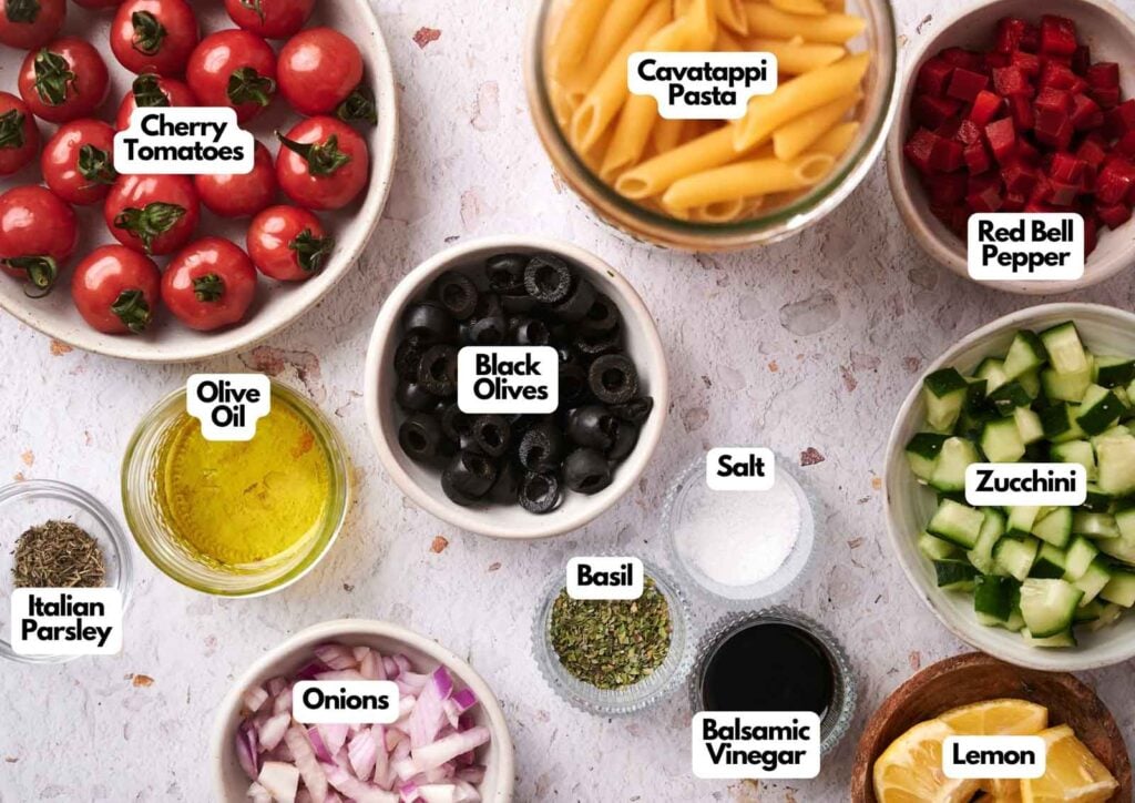 A colorful selection of pasta salad ingredients, including cherry tomatoes, cavatappi pasta, red bell pepper, zucchini, black olives, onions, olive oil, salt, basil, Italian parsley, balsamic vinegar, and lemon.