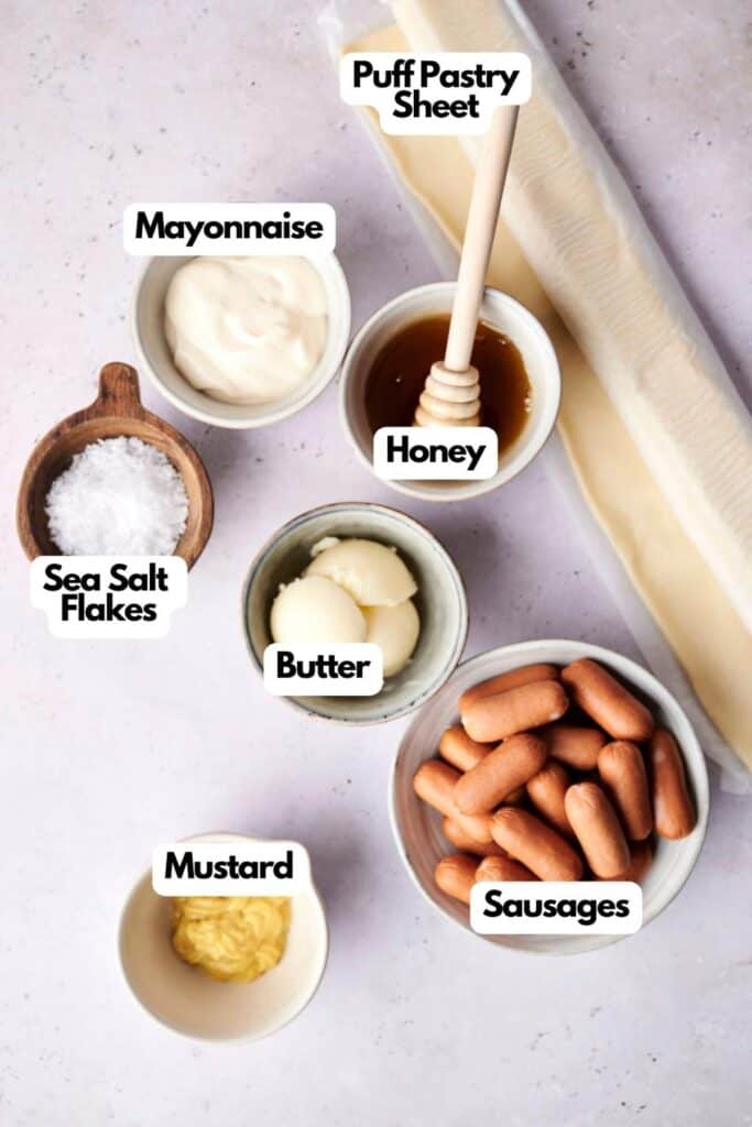Ingredients for a recipe including a puff pastry sheet, honey, sausages (perfect for making Pigs in a Blanket), mayonnaise, butter, mustard, and sea salt flakes arranged on a white surface.