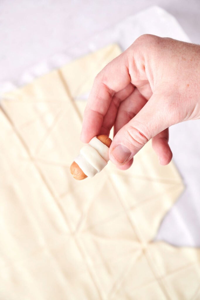 A hand holding a small sausage wrapped in dough, resembling classic Pigs in a Blanket, with a sheet of dough in the background on a flat surface.