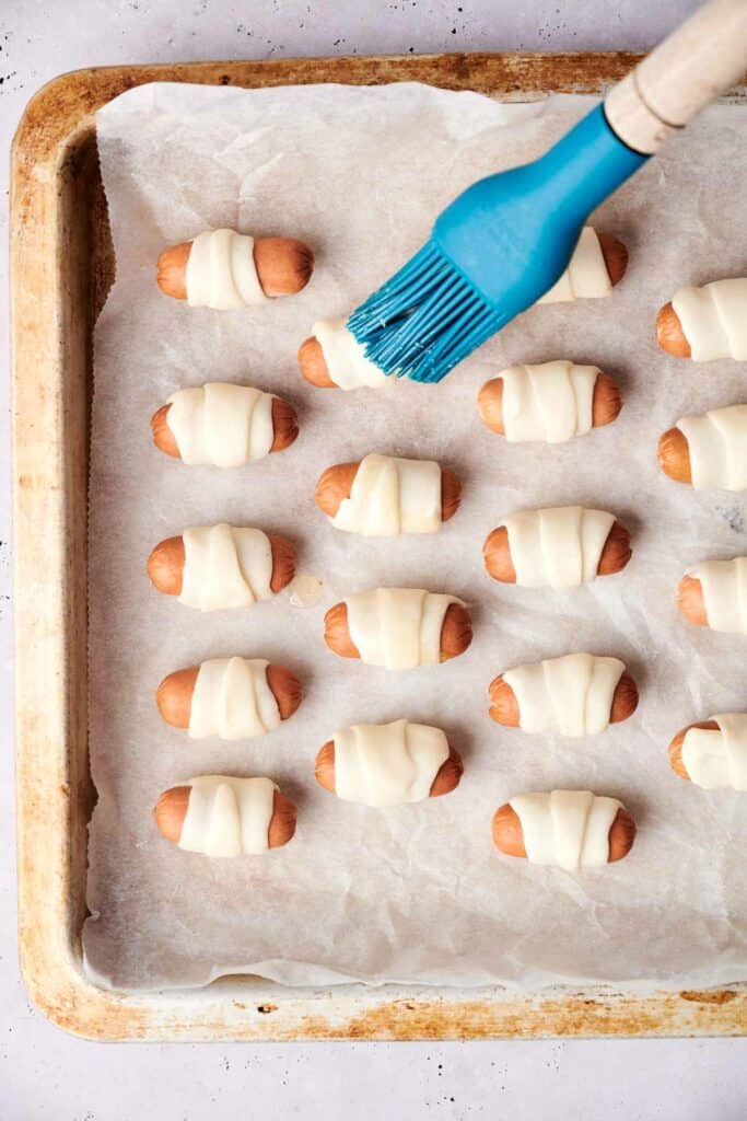 A baking tray filled with uncooked pigs in a blanket is set for preparation. Someone is meticulously brushing the delightful pigs in a blanket with egg wash using a blue brush.