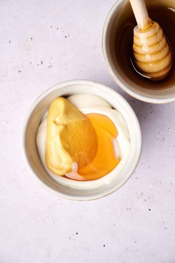 A bowl of yogurt topped with honey and a dollop of mustard, next to a small dish of honey with a honey dipper—perfect for pairing with pigs in a blanket.