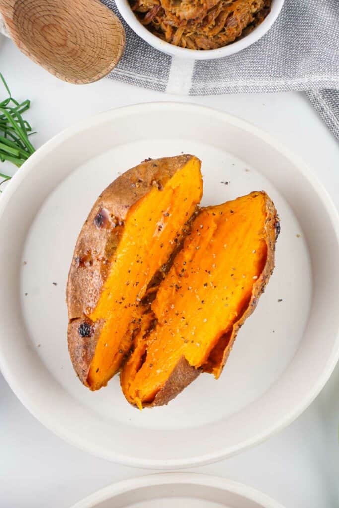 A baked sweet potato split into two halves, served on a white plate. The flesh is bright orange and sprinkled with black pepper.