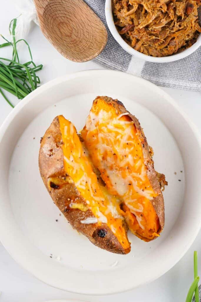 A baked potato on a white plate, split open and topped with melted cheese. A wooden spoon and a bowl of shredded meat are visible in the background.