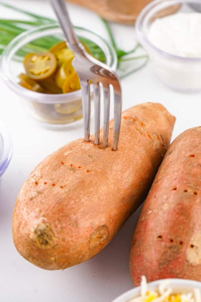 A fork piercing a raw sweet potato, surrounded by bowls of ingredients including sliced jalapeños, cheese, and sour cream on a white surface.
