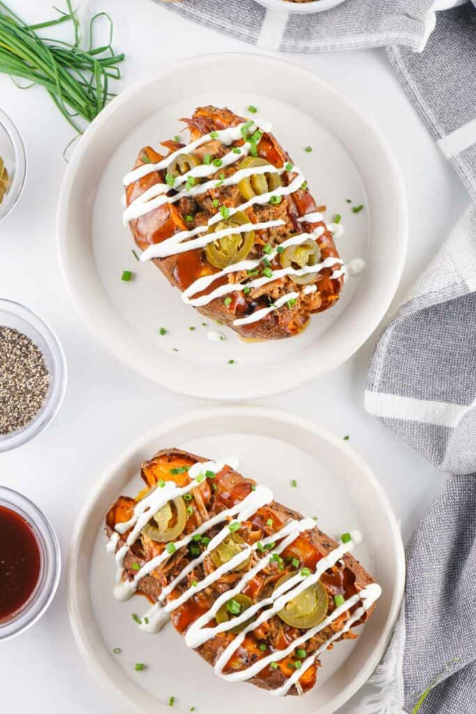 Two plates of baked sweet potatoes topped with sour cream, pickles, and chives, placed on a white table with a gray-striped napkin and small bowls of condiments nearby.
