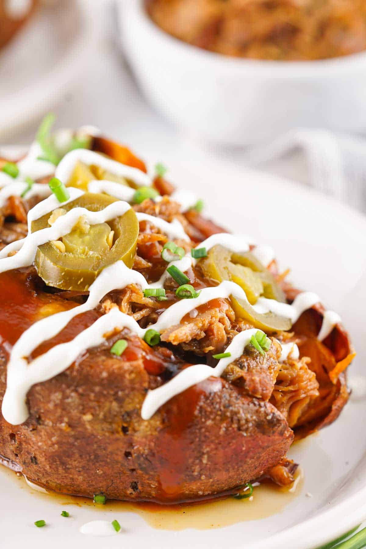 A pulled pork baked potato topped with shredded meat, jalapenos, chives, barbecue sauce, and sour cream is served on a white plate. A bowl with a similar dish is visible in the background.