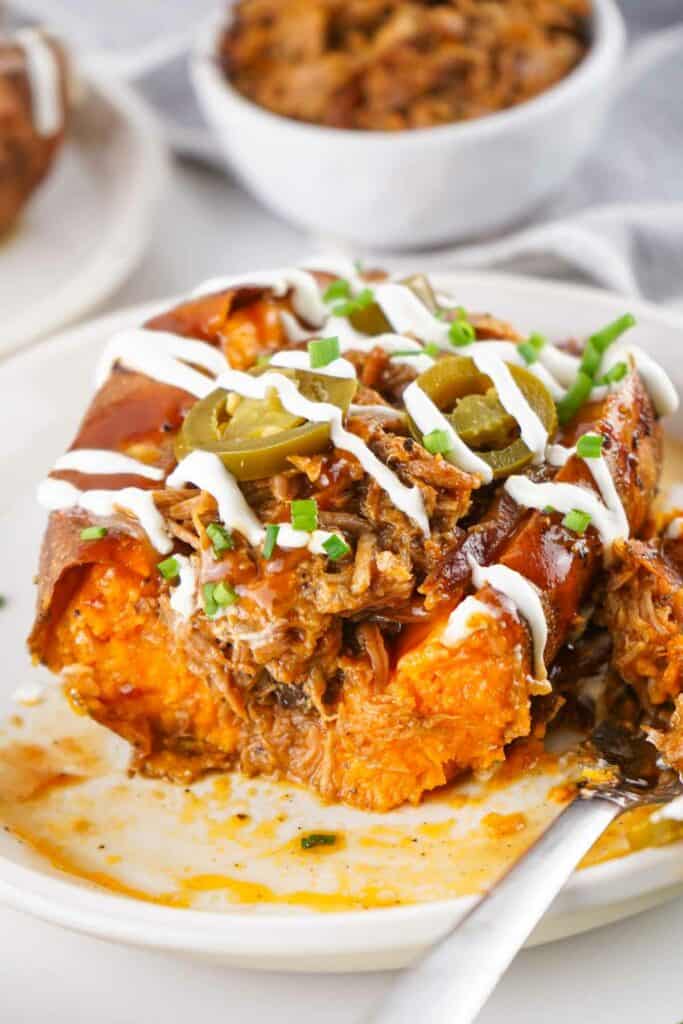 A baked sweet pulled pork potato topped with shredded meat, sour cream, jalapenos, and chopped chives on a white plate. A fork is partially shown on the side.