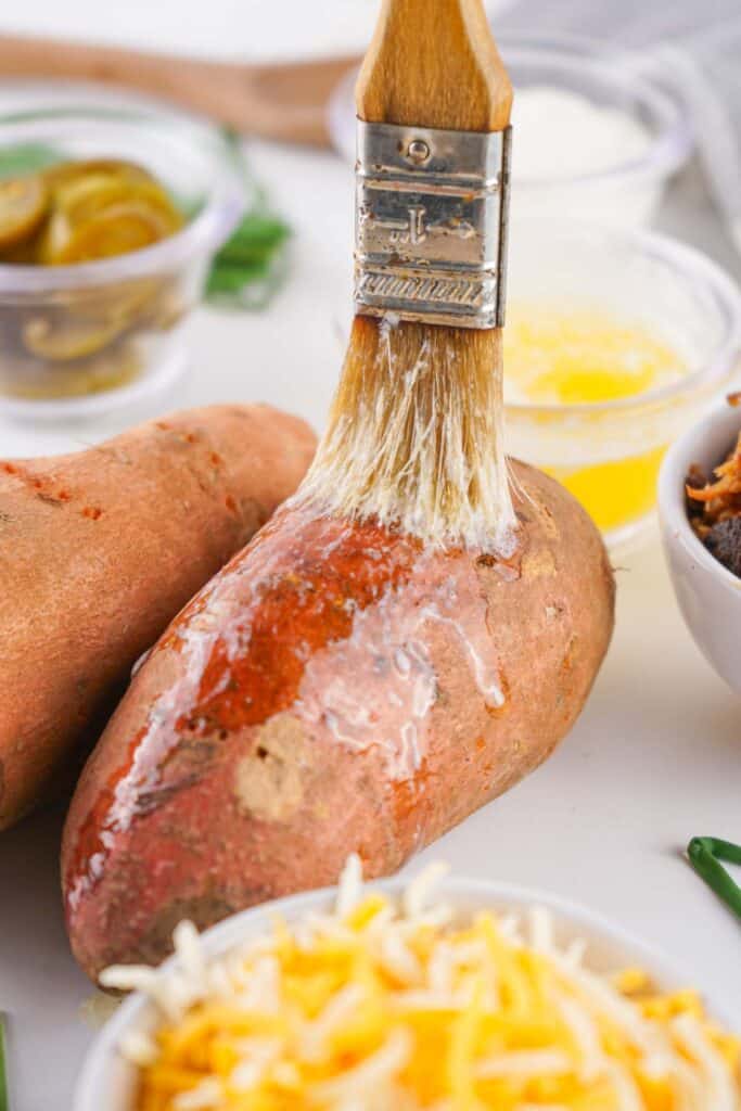 A brush applies melted butter to a raw sweet potato. Cups of grated cheese, jalape&ntilde;os, and other ingredients are in the background.