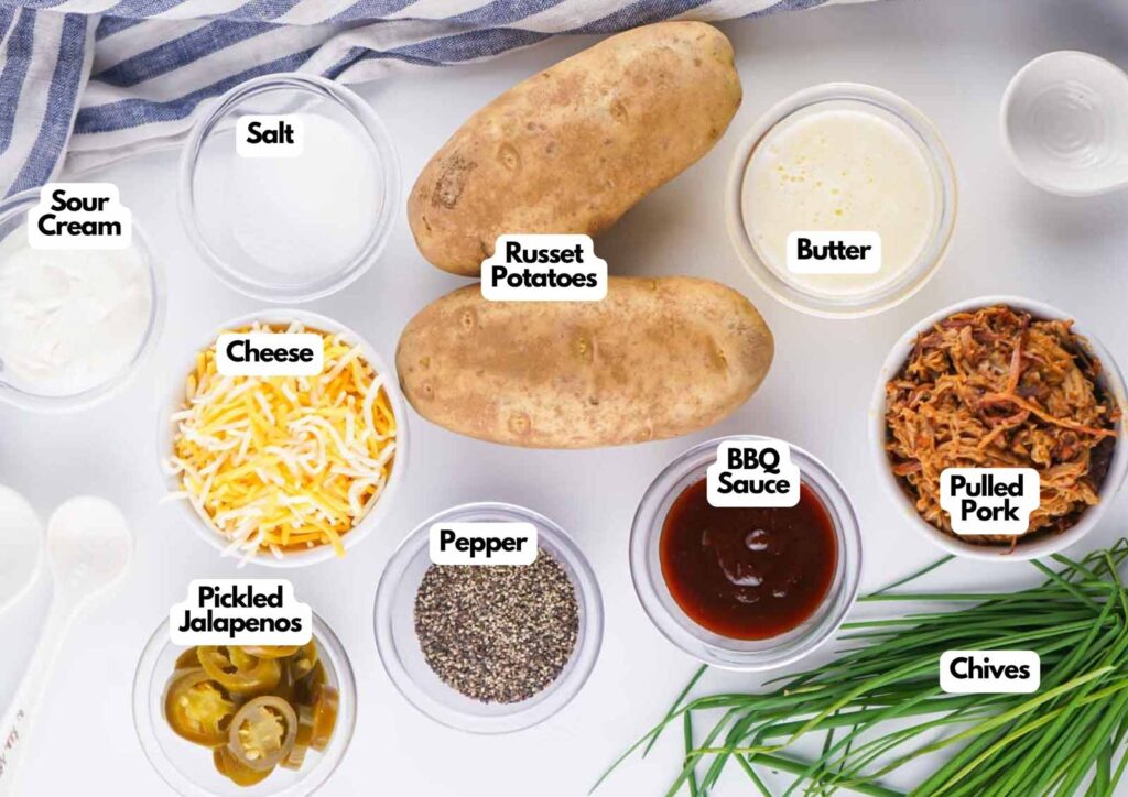 Ingredients for pulled pork potatoes are arranged on a white surface, including russet potatoes, tender pulled pork, cheese, chives, sour cream, pickled jalapeños, butter, BBQ sauce, salt, and pepper.