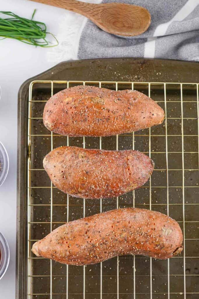 Three seasoned sweet potatoes on a wire rack over a baking sheet, with a wooden spoon and towel in the background.