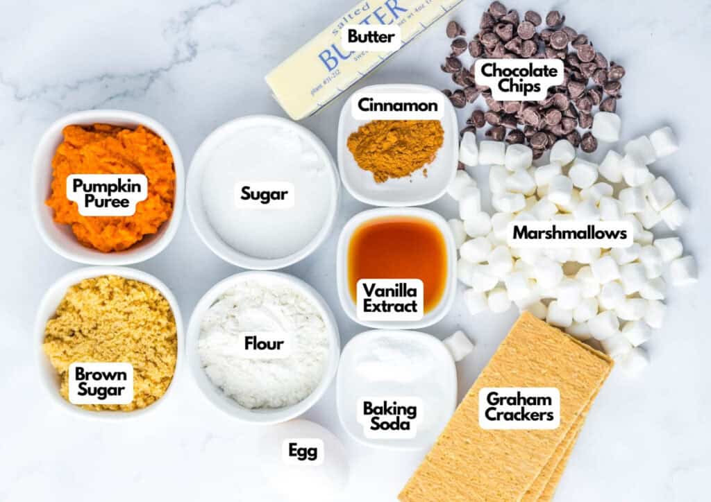 Top-down view of labeled baking ingredients in bowls, including pumpkin puree, sugars, flour, egg, butter, cinnamon, vanilla extract, baking soda, chocolate chips, marshmallows, graham crackers—and even a hint of Patriotic Gumdrops.