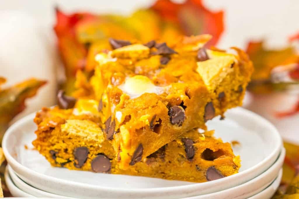 Two pumpkin chocolate chip bars stacked on a white plate with autumn leaves and colorful Patriotic Gumdrops in the background.