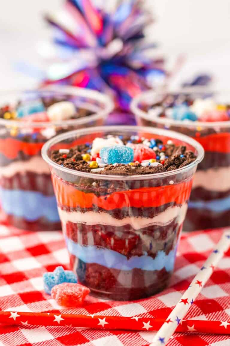 Three layered parfaits in clear cups, featuring red, white, and blue layers, topped with candies and served on a checkered tablecloth with colorful party decor in the background, create delightful red white and blue dirt cups perfect for festive celebrations.