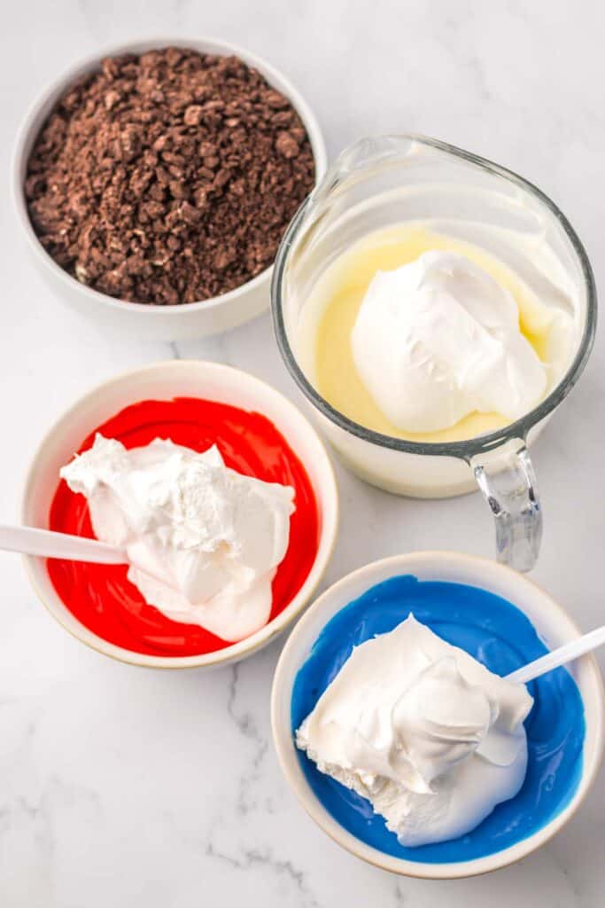 A bowl of chocolate crumbles, a jug of white liquid with a dollop of cream, two bowls containing red and blue liquids each with a white dollop, reminiscent of red white and blue dirtcups, and a spoon on a white marble surface.