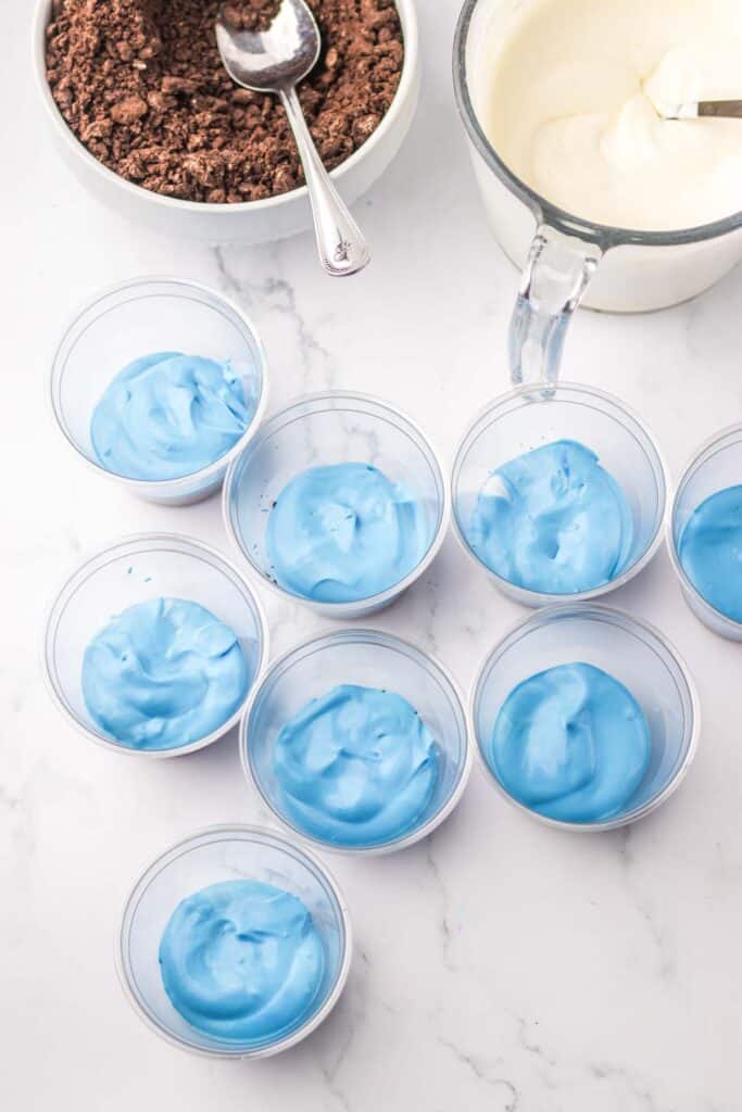 Several clear plastic cups filled with blue whipped cream, part of a delightful red white and blue dirtcups display, are arranged on a marble surface. Nearby are a bowl of chocolate crumbs with a spoon and a pitcher of white whipped cream.