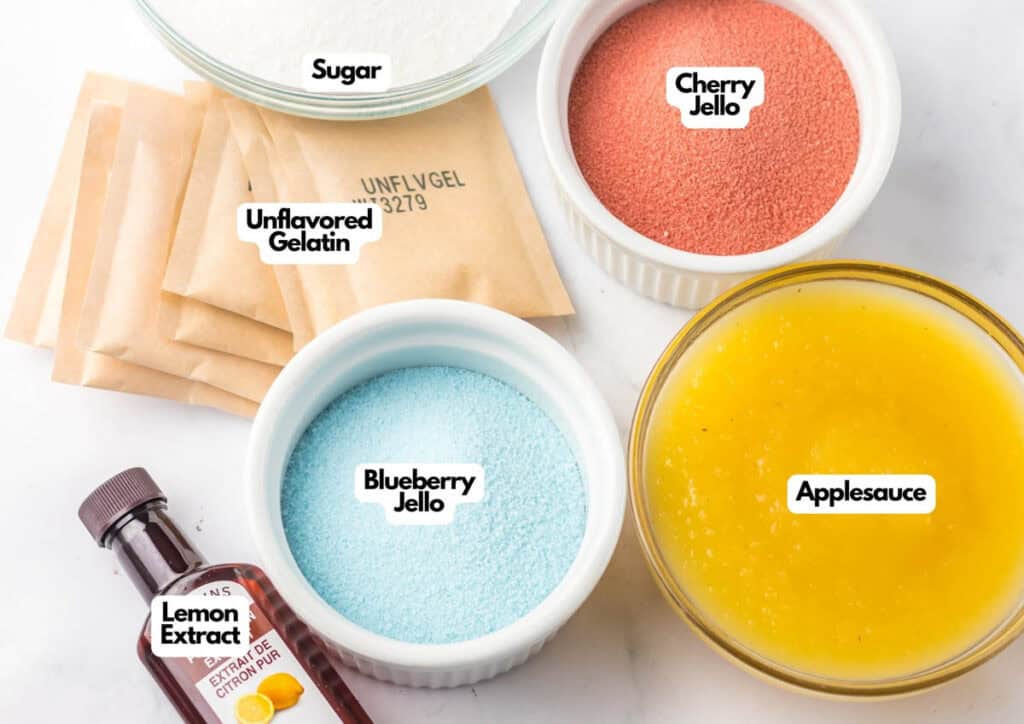 Various ingredients are arranged on a counter for making Patriotic Gumdrops, including bowls of sugar, cherry jello, blueberry jello, applesauce, packets of unflavored gelatin, and a bottle of lemon extract.