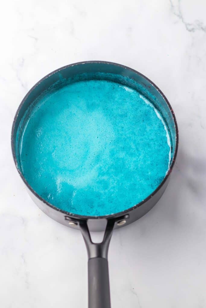 A black saucepan filled with bright blue liquid sits on a white marble surface.