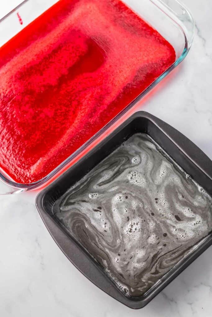 Two baking dishes on a marble countertop: one with a red gelatin mixture and the other with a swirled black gelatin mixture.