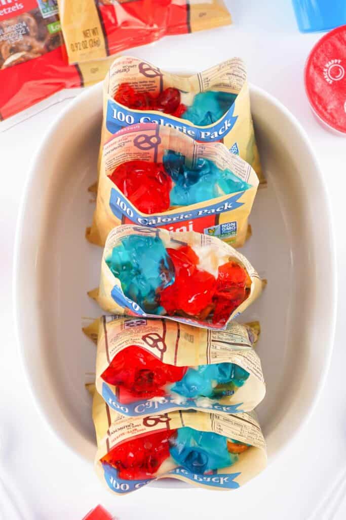 A white tray holds several 100-calorie snack packs of mini pretzels, each topped with red and blue jelly, perfect for a Red White and Blue Walking Pretzel Salad.