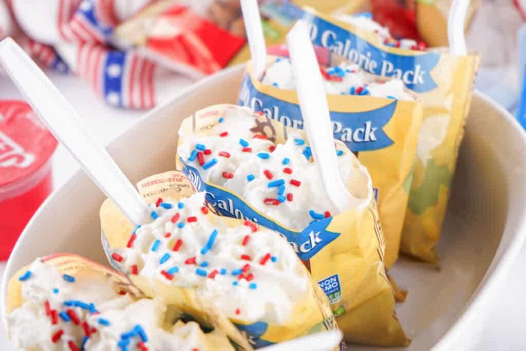 Packets of 100 calorie popcorn topped with whipped cream, red, white, and blue sprinkles, and a few Patriotic Gumdrops, each served with a white plastic spoon in a white dish.