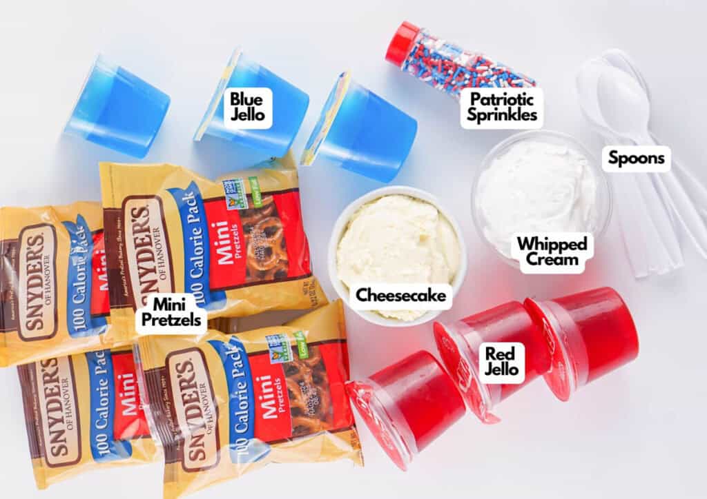 Assorted ingredients on a white surface: blue and red jello cups, mini pretzels, whipped cream, cheesecake, patriotic sprinkles, Patriotic Gumdrops, and plastic spoons.