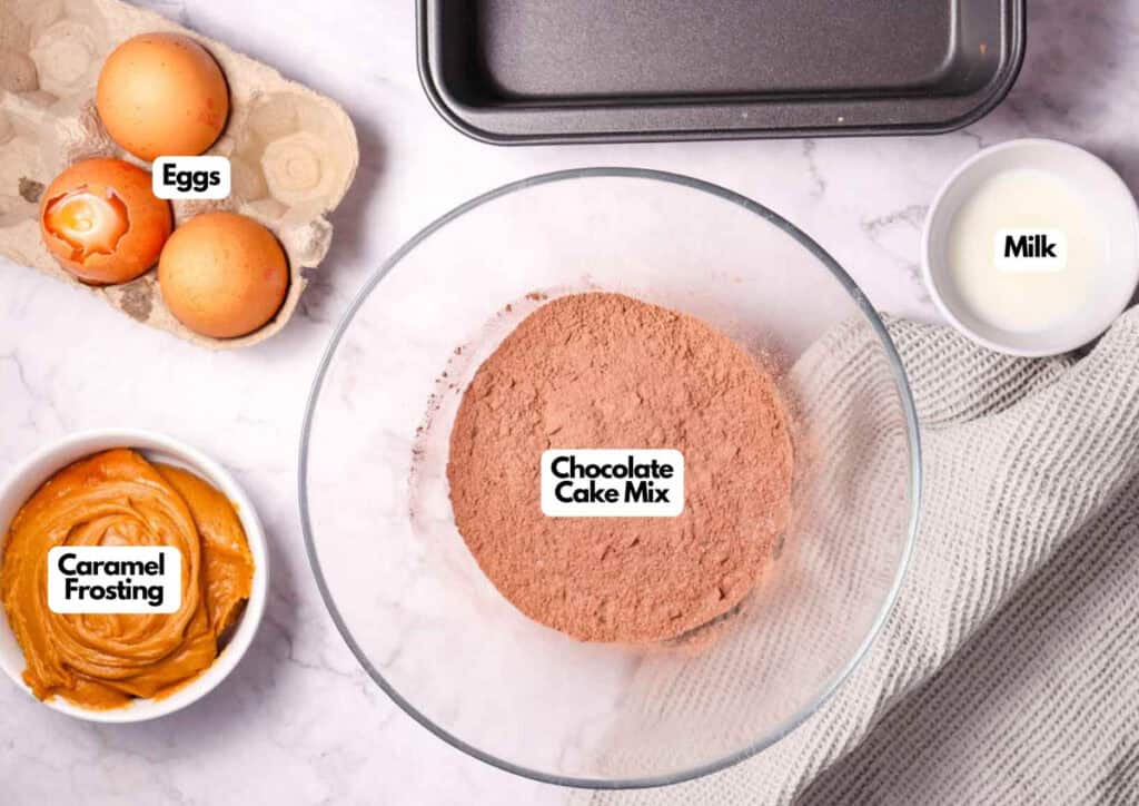 A bowl of chocolate cake mix, a container of caramel frosting, three eggs, a cup of milk, Patriotic Gumdrops, and a baking pan are arranged on a marble countertop.