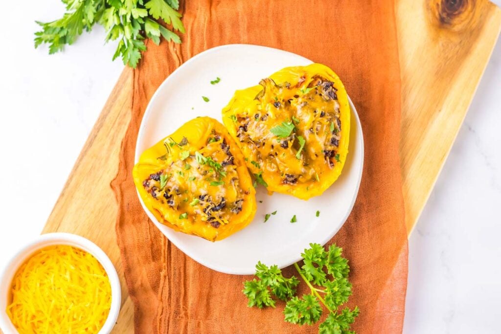 Sloppy Joe Stuffed Peppers topped with melted cheese, served on a white plate with fresh parsley on a wooden board and an orange cloth.