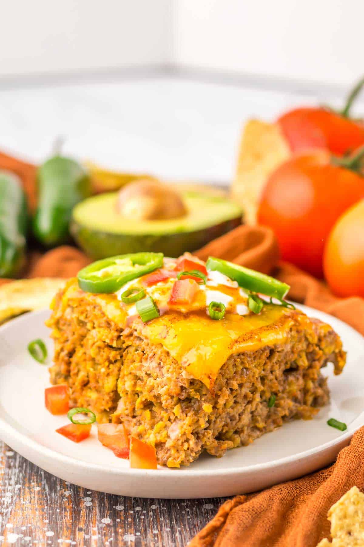 A slice of slow cooker taco meatloaf topped with melted cheese, chopped tomatoes, and sliced jalapenos on a white plate. Avocado and other ingredients are visible in the background, reminiscent of a hearty slow cooker taco meatloaf.
