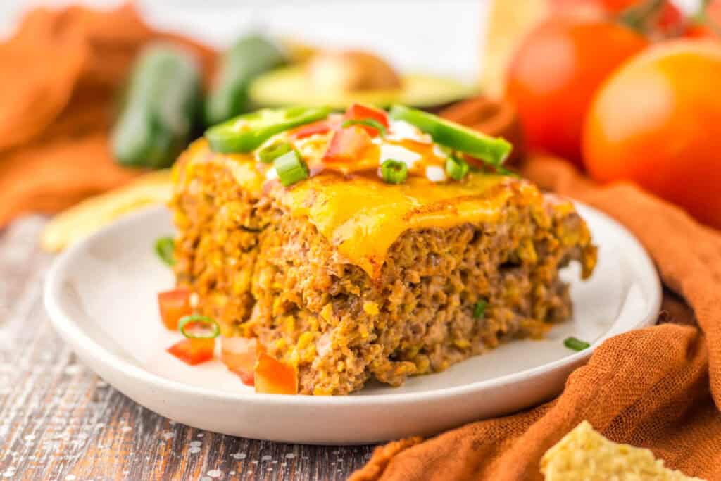 A slice of slow cooker taco meatloaf, topped with melted cheese, tomatoes, and jalapenos on a white plate. Fresh vegetables are in the background.