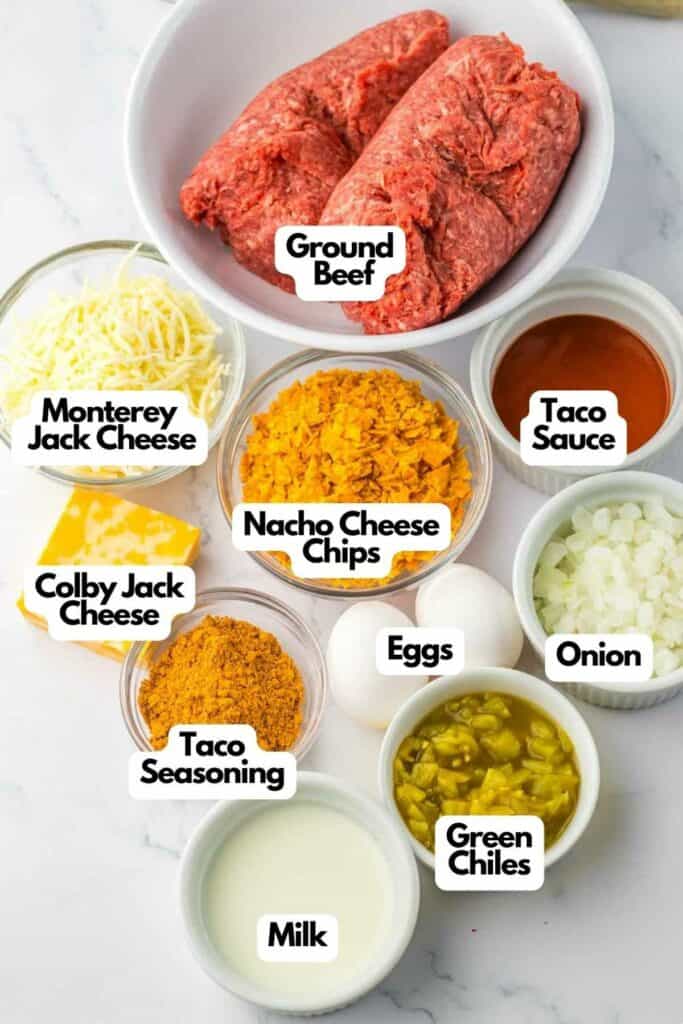 Ingredients for a slow cooker taco meatloaf recipe arranged on a white surface, including ground beef, Monterey Jack cheese, taco sauce, Colby Jack cheese, nacho cheese chips, taco seasoning, eggs, onion, green chilies, and milk.