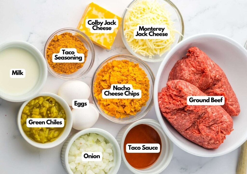 Ingredients for slow cooker taco meatloaf arranged on a counter: ground beef, eggs, milk, taco seasoning, green chiles, onion, taco sauce, nacho cheese chips, Colby Jack cheese, and Monterey Jack cheese.
