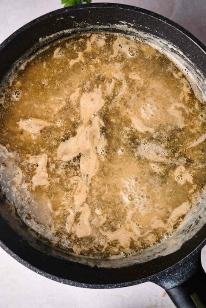 A large black pot filled with simmering broth, pieces of cooked chicken, and a few Swedish meatballs. The liquid appears to be seasoned, with bubbles and frothy patches on the surface.