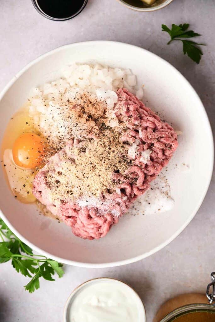 A bowl contains raw ground meat, a cracked egg, chopped onions, and seasonings—perfect beginnings for Swedish meatballs. Fresh parsley and additional ingredients are visible around the bowl.