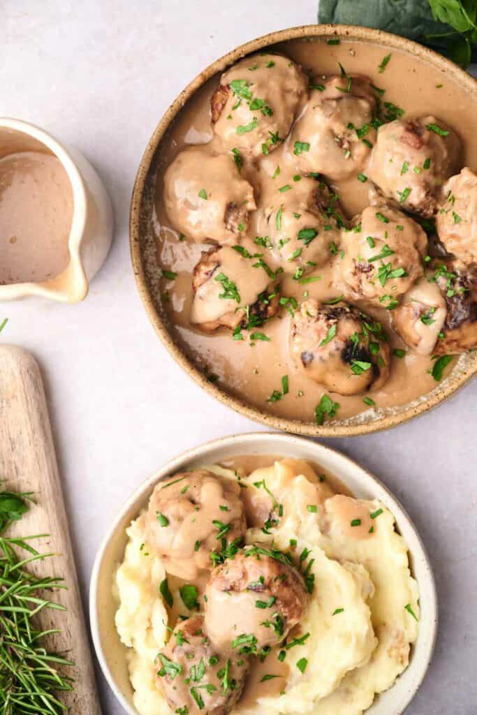 A bowl of Swedish meatballs covered in gravy sits next to a serving of mashed potatoes topped with meatballs and gravy. A sprig of rosemary and a jug of gravy are beside the bowls.