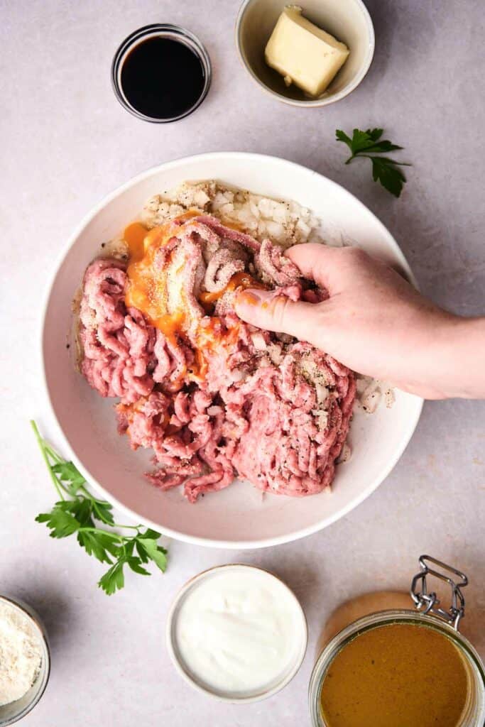 A hand mixing ground meat, egg yolk, and spices in a white bowl, preparing mouthwatering Swedish meatballs, surrounded by small bowls containing sauce, flour, butter, and other essential ingredients.
