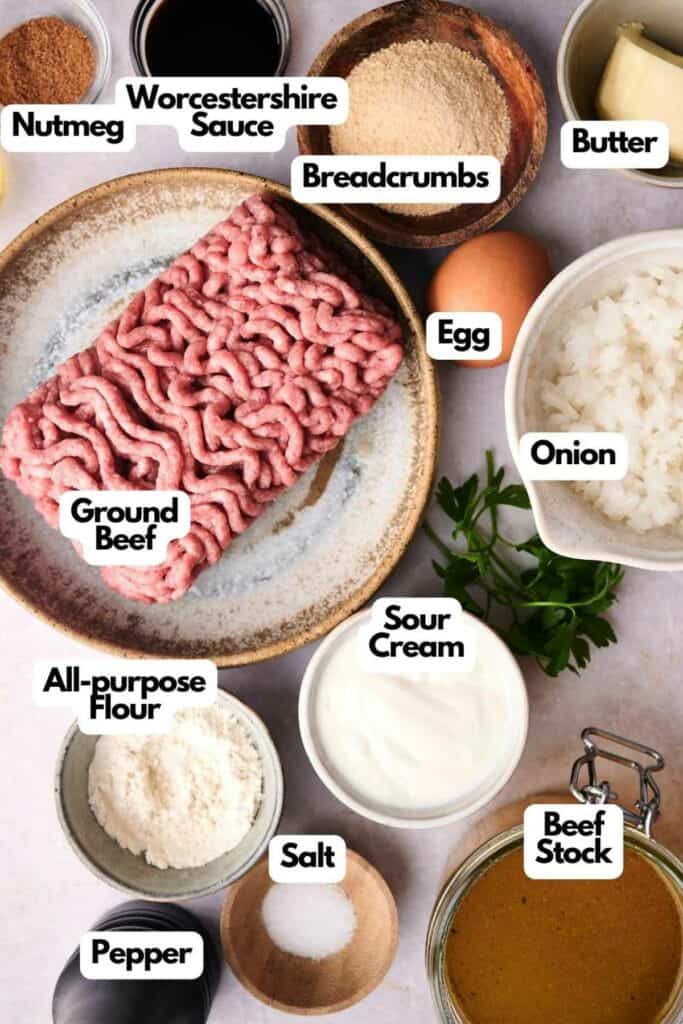 Ingredients for Swedish meatballs arranged on a surface, including ground beef, breadcrumbs, butter, egg, onion, sour cream, Worcestershire sauce, nutmeg, all-purpose flour, pepper, salt, and beef stock.