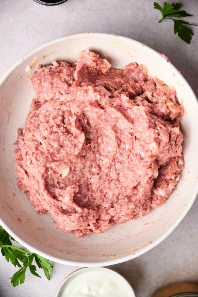 A close-up of a bowl filled with a mixture of raw ground meat, likely being prepared for a recipe like Swedish meatballs. Some parsley leaves are visible around the bowl.