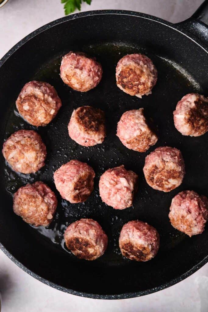 A black frying pan contains several partially cooked Swedish meatballs, some browned on one side.