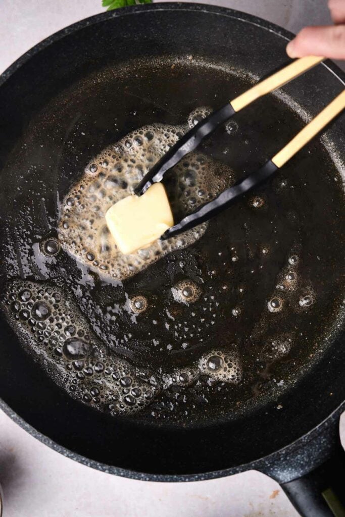 A cube of butter is being held by black and beige kitchen tongs over a bubbling liquid in a black frying pan, preparing the base for savory Swedish meatballs.