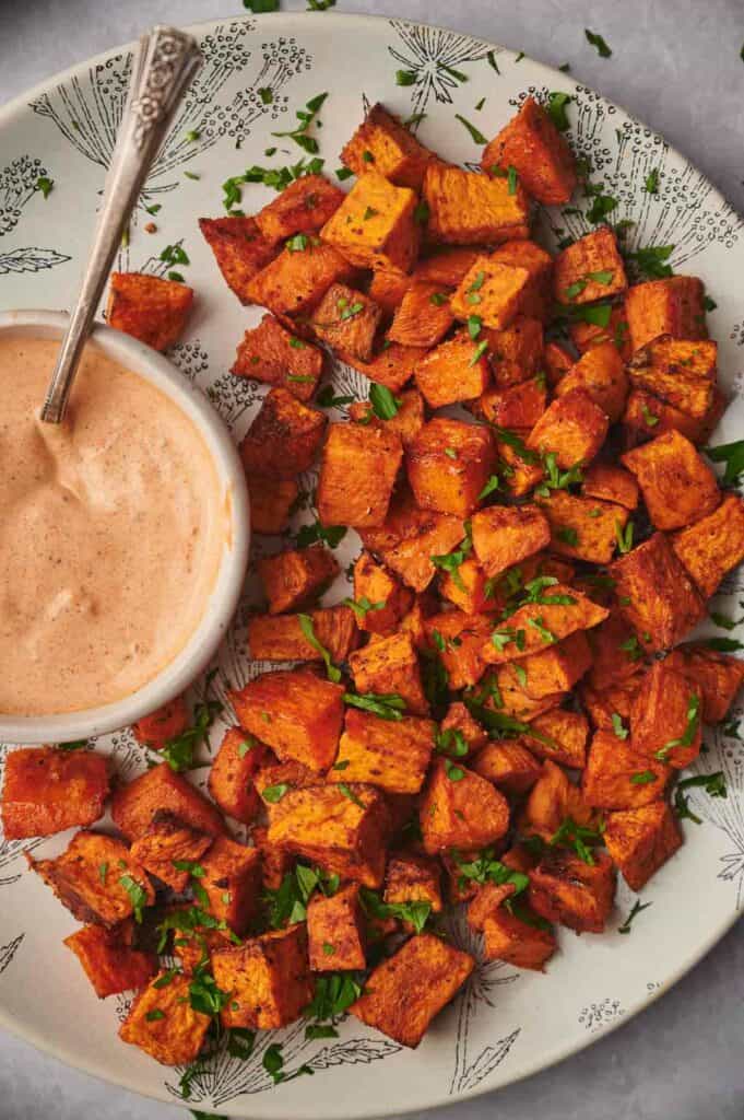 A plate of seasoned, cooked sweet potato cubes garnished with chopped herbs, served alongside a small bowl of dipping sauce with a spoon.