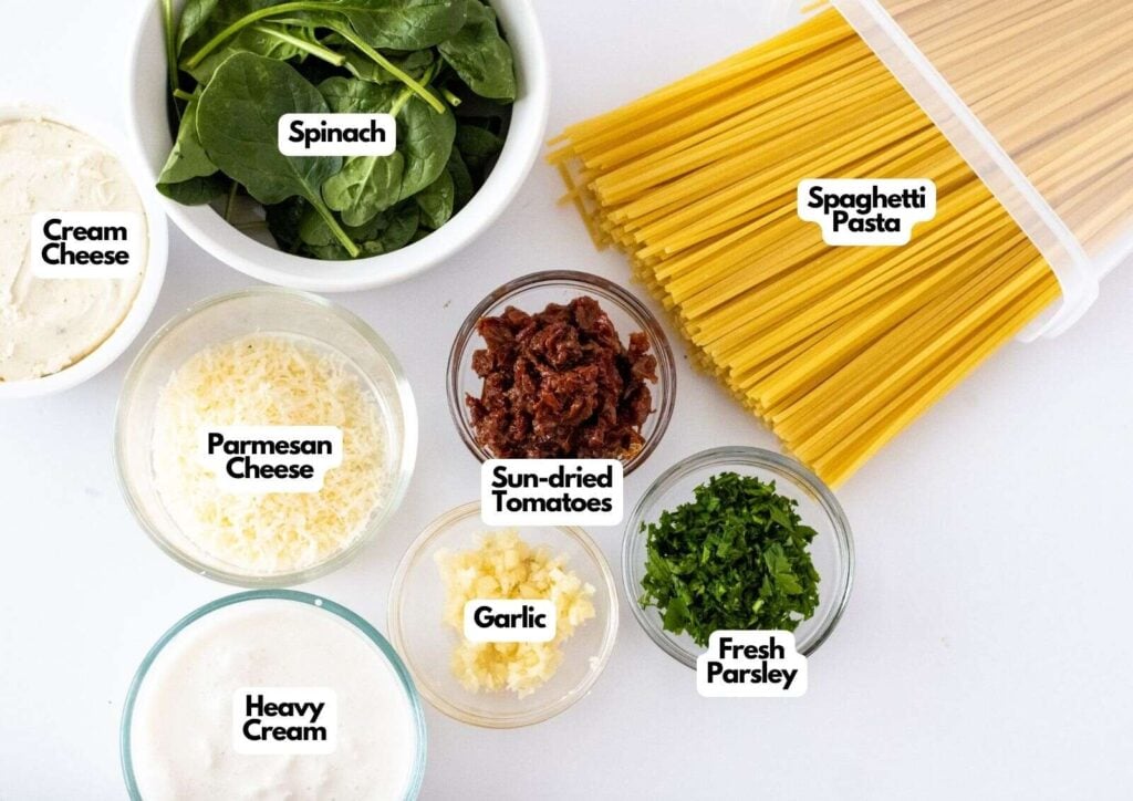 Ingredients for a Creamy Tuscan Pasta dish are arranged on a white surface: spinach, spaghetti pasta, cream cheese, parmesan cheese, sun-dried tomatoes, garlic, fresh parsley, and heavy cream.
