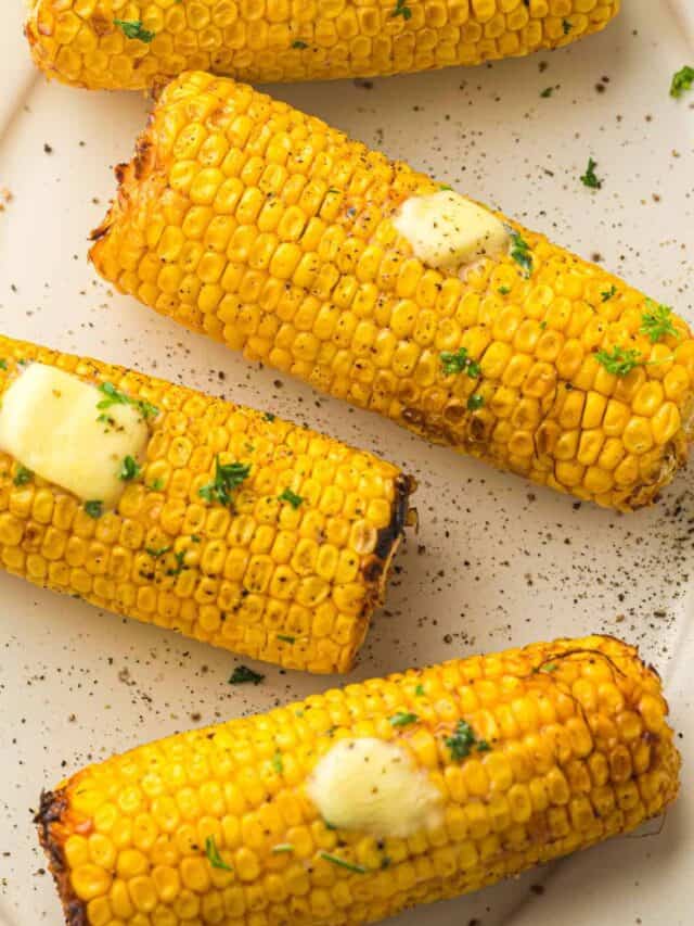 Four pieces of grilled corn on the cob topped with melted butter and garnished with chopped herbs on a white plate.