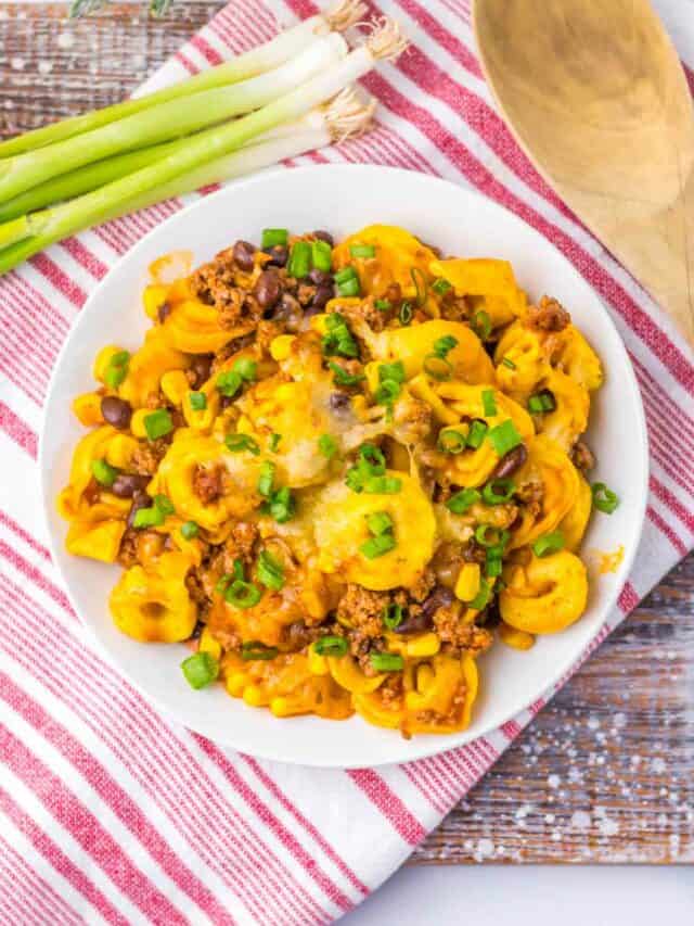 A white dish filled with cheddar cheese pasta, black beans, corn, ground meat, and green onions sits on a red and white striped cloth next to a wooden spoon and bunch of green onions.