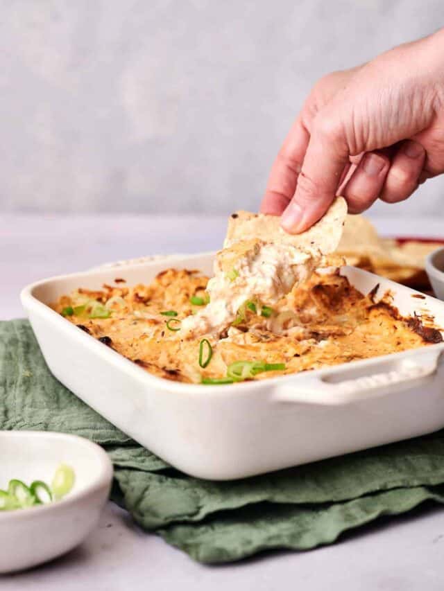 A hand is dipping a tortilla chip into a cheesy baked dip in a white dish, garnished with green onions.