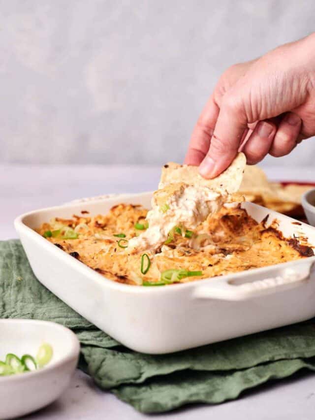 A person's hand is dipping a tortilla chip into a baked cheese dip topped with green onions in a white baking dish.
