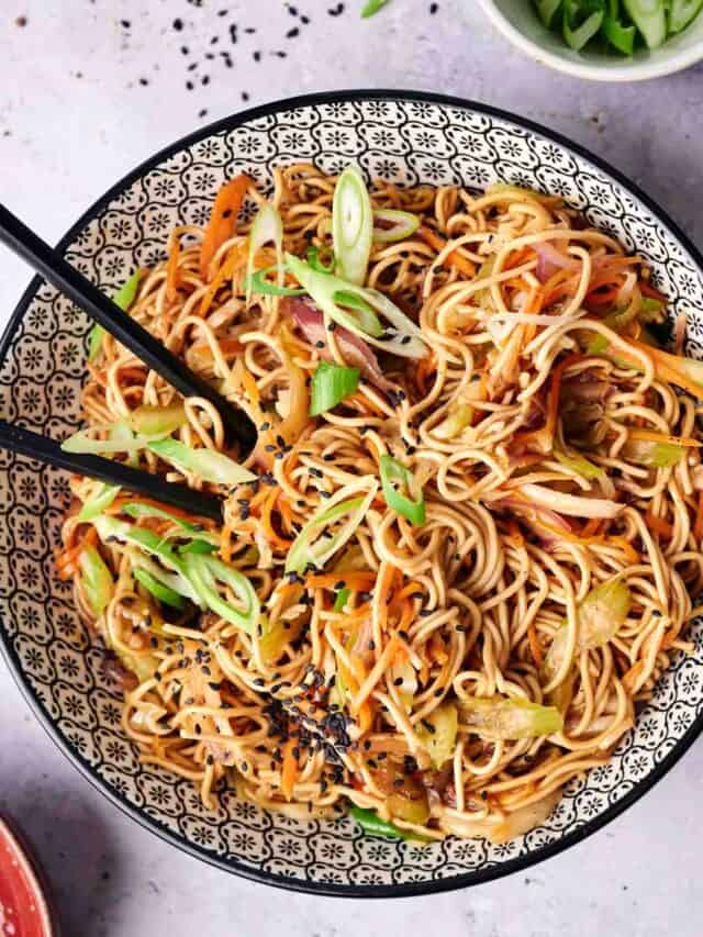 A bowl of mixed vegetable lo mein garnished with green onions and sesame seeds, with a pair of black chopsticks resting on the edge.