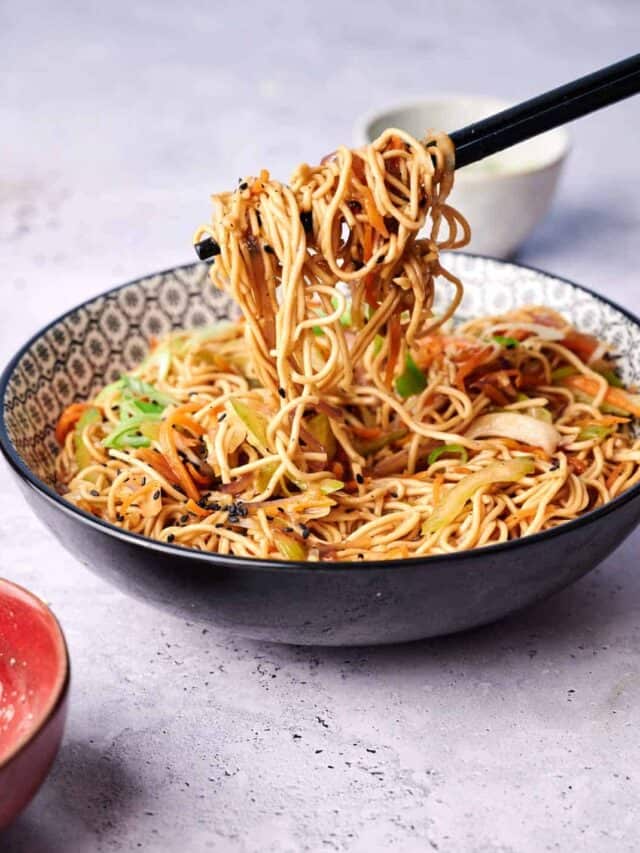 Bowl of stir-fried noodles with vegetables, shown with chopsticks lifting a portion.