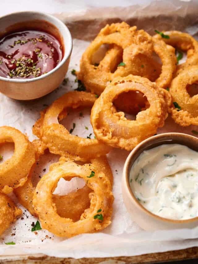 A serving of crispy fried onion rings is arranged on a parchment-lined tray, accompanied by two dipping sauces—one red with herbs and one white with green flecks.