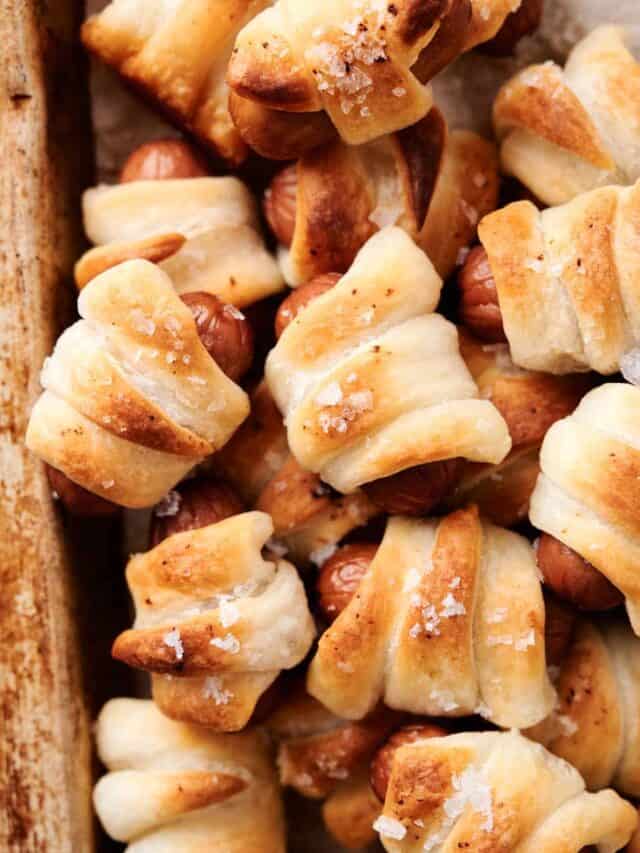 Close-up of pastry-wrapped sausages, baked to a golden brown with a sprinkle of coarse salt on top. The pastries are arranged tightly on a baking sheet.